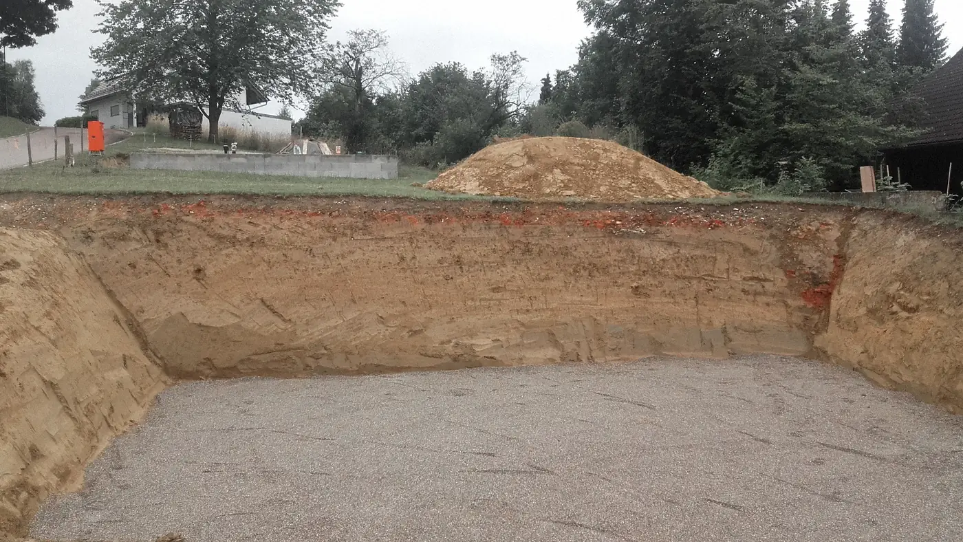 Fouille terminée pour une maison : talus de terre et fond garni de gravier, tas de déblais au fond